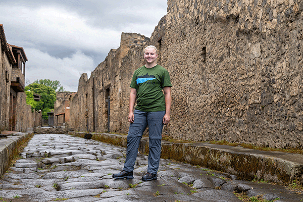 A female student standing on a rock road and looking at the camera while posing for a photo in the streets of the ancient city of Pompeii in Italy. Stone ruins of building are on each side of the roadway.