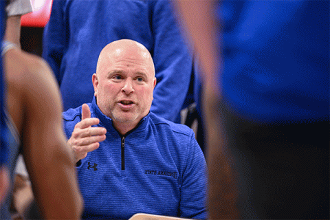 Middle-aged white male wearing a blue zipped-up shirt and talking to basketball players with their back to the camera.